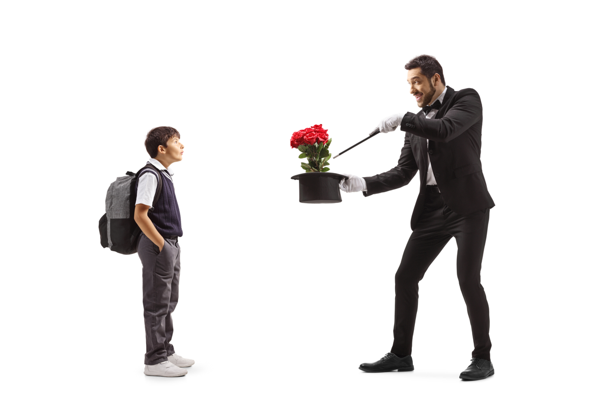 Image of a magician making flowers appear out of a top hat via his wand, performing directly for an impressed little boy wearing a large backpack.