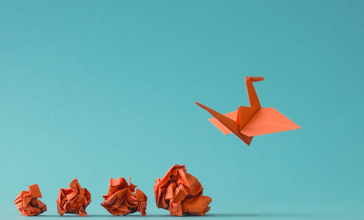 Photograph of a series of crumpled orange balls of paper and an orange folded crane flying in the air, against a turquoise background.