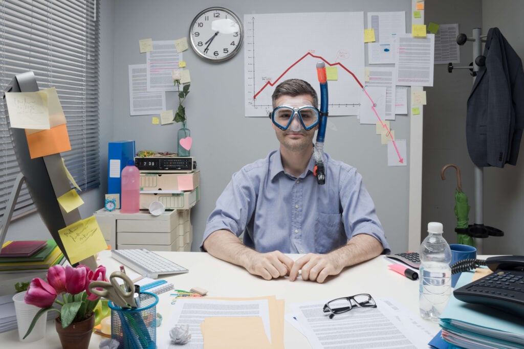 Image of a man in an office wearing a snorkel and goggles, with a chart beyond him showing the numbers going down so much they have to extend it with post-it notes.