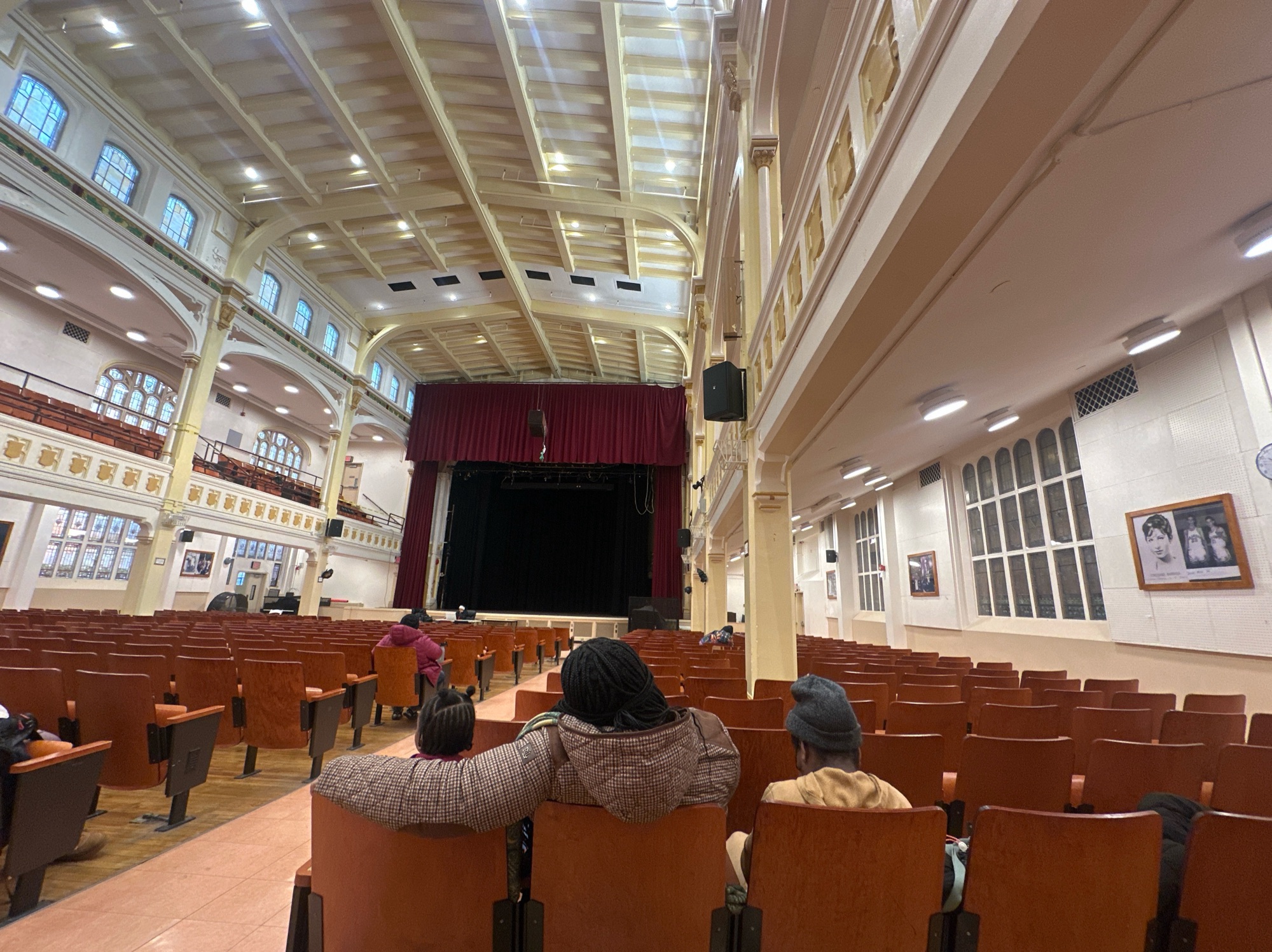 A somewhat ornate auditorium. People sitting. On the right a framed photo of Barbra Streisand.