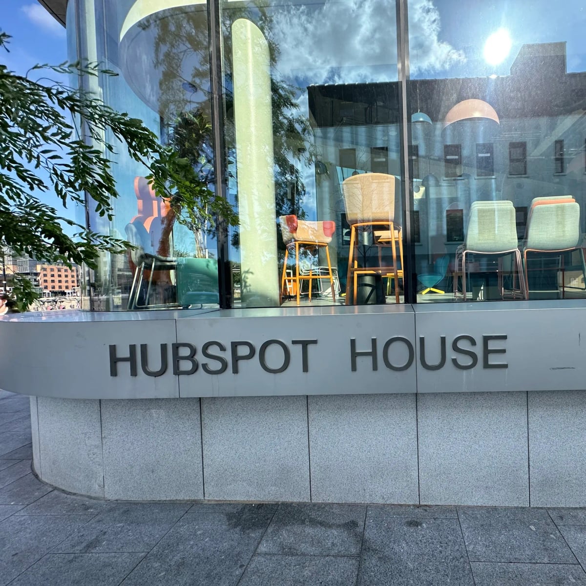 Exterior shot. A ground-level glassed in modern lobby. Lots of chairs. Metal lettering reads HUBSPOT HOUSE.