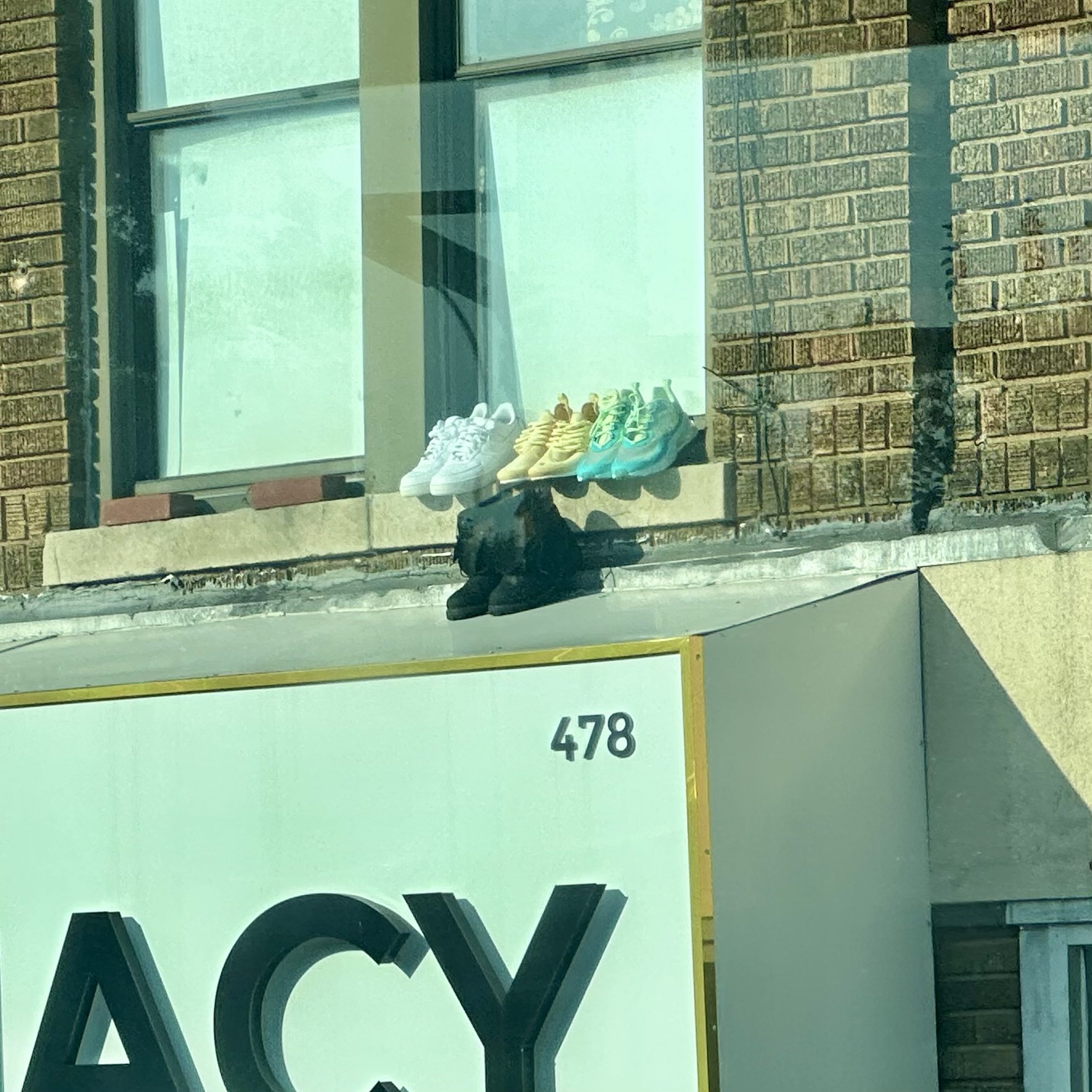 Boots and three pairs of sneakers on a window ledge. The sneakers are white, yellow, and turquoise. The boots are black and are placed right on the metal awning of the pharmacy below.