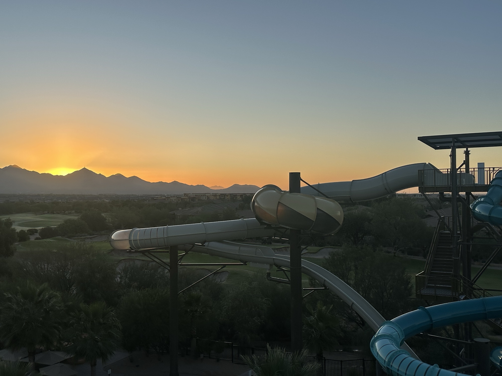 The sun rises over distant mountains—a beautiful desert scene. In the darker foreground a large looping water slide fills in half the frame on the right.
