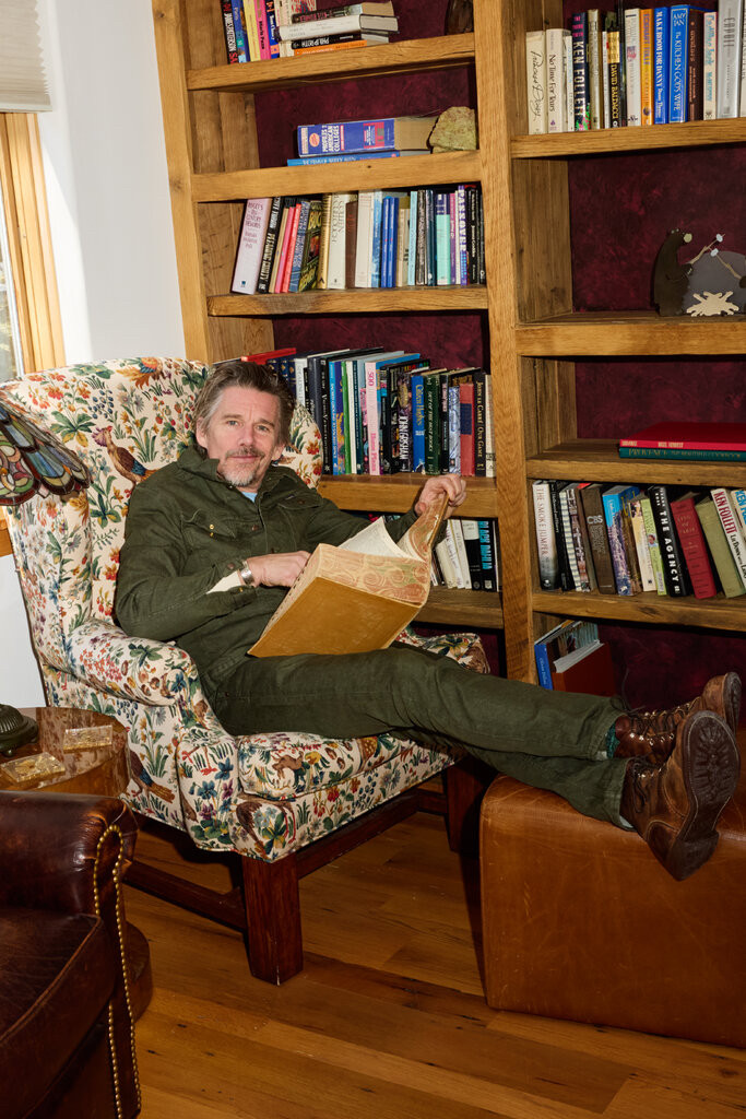 A smirking Ethan Hawke dressed all in military shades of green with boots on, in a hideous armchair upholstered with a bird and plants print, one of those things that is so relentlessly ugly it signifies sophistication and probably costs $9,000, in front of a bunch of books. He's reading a massive, marble-edged, leather-bound book that appears to be a dictionary. He pulls it off because he's just an earnest poetic sex angel and we all know it but literally anyone else would be dragged for eons through every tranche of hell.