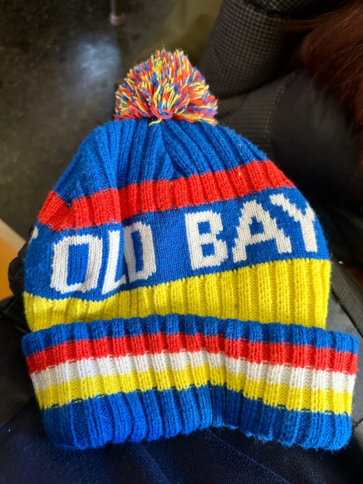 A mass-produced knit cap in extremely bright blue, yellow, white, and red stripes, with a large blue, yellow, and red pom-pom on top. The forehead-covering part of the cap reads, in enormous white knit-pixelated letters, OLD BAY.